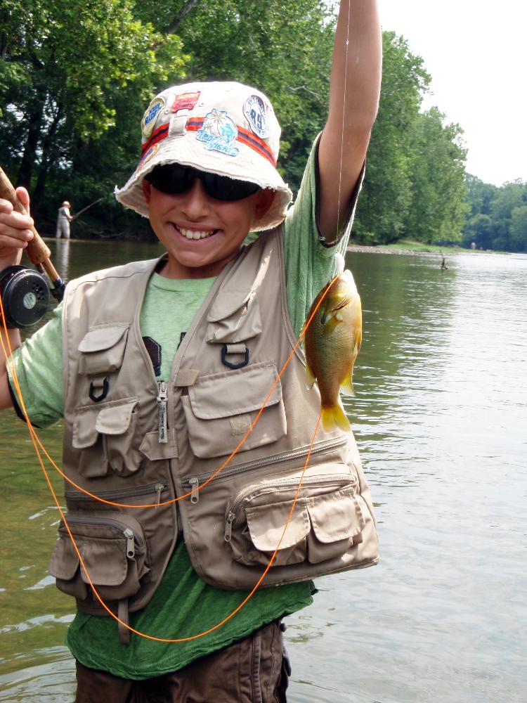 Young boy holding a redbreast sunfish he caught using a fly rod.  He is wearing a fishing vest, sunglasses a cool hat and a huge smile while standing in a clear Shenandoah River