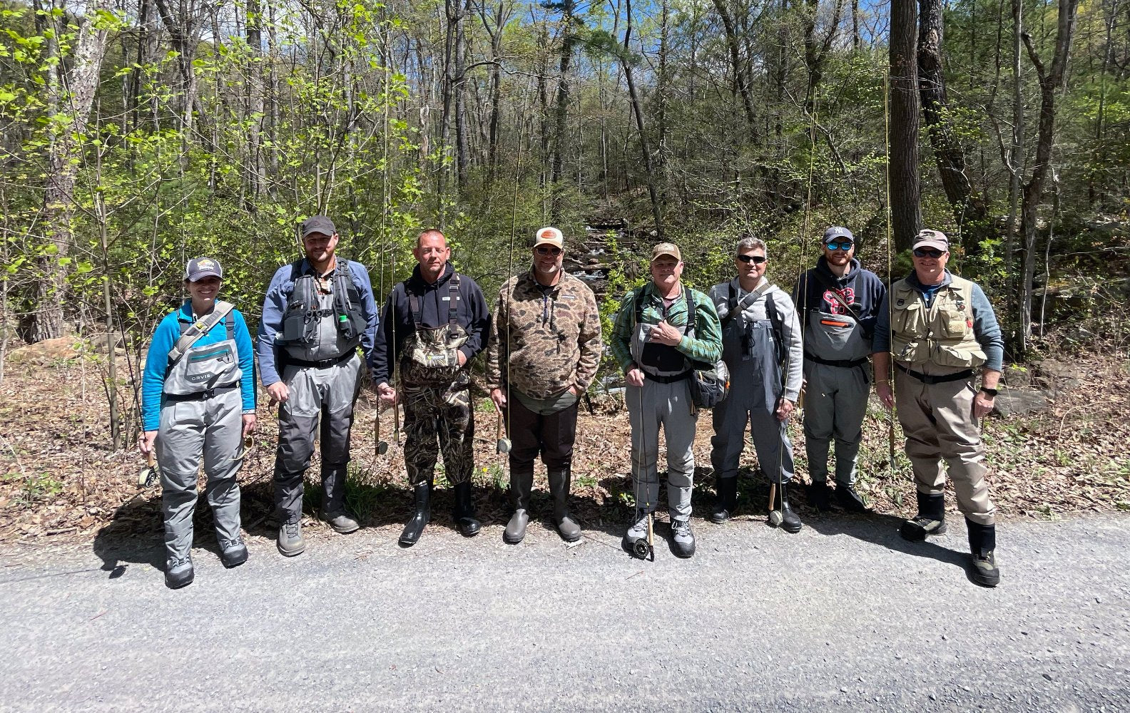 Mountain Trout On The Stream Fly Fishing School showing several anglers dressed in waders, vests and holding fly rods.  They are surrounded by trees and a small mountain brook is in the background