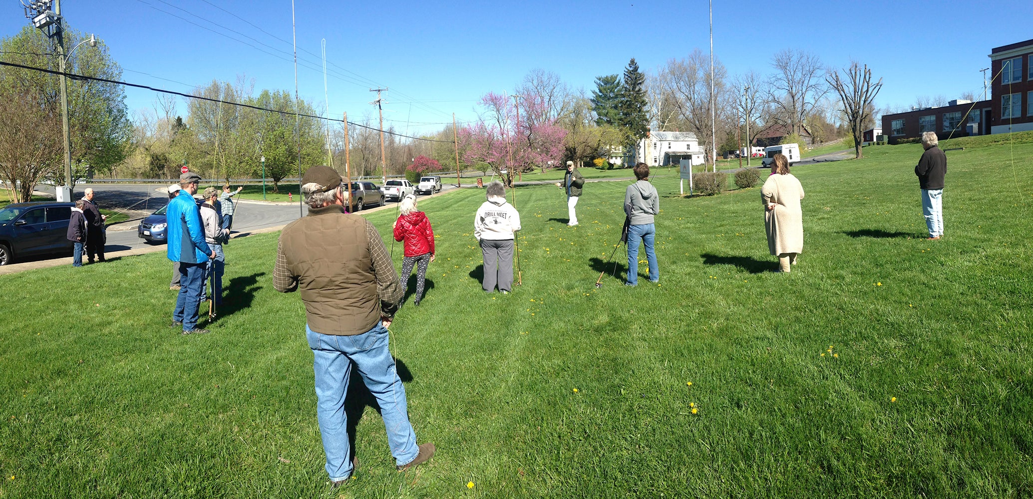 People are lined up on our fly casting lawn taking a lesson from Harry Murray.  They are spread across the green grass from left to right.  Each of them are holding a fly rod.