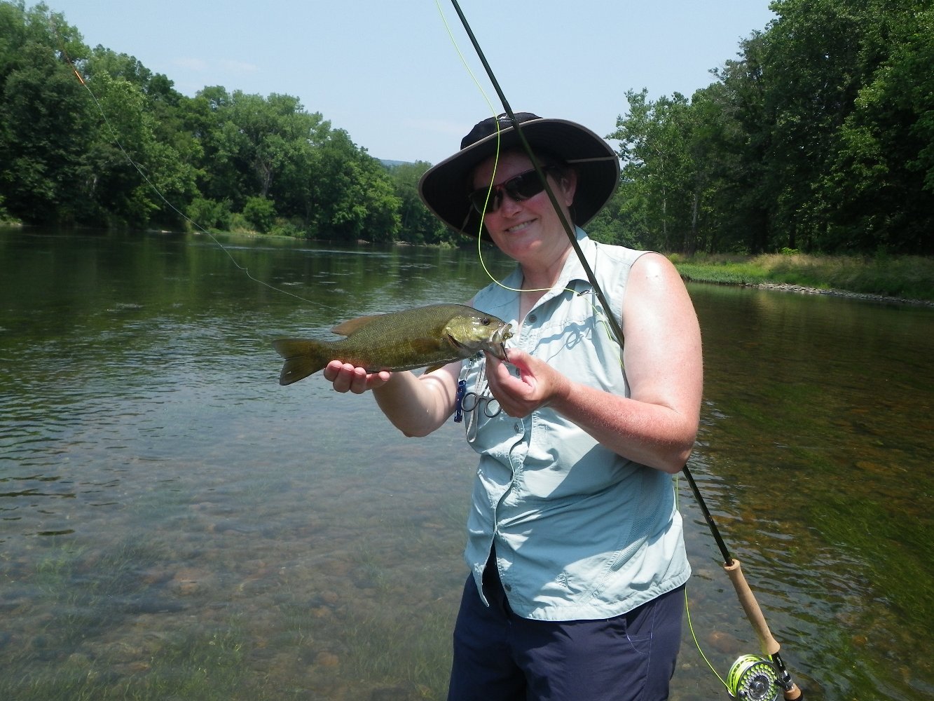 Lady wearing a hat holding a smallmouth bass and a fly rod while standing in a clear river in Virginia - Shenandoah River with green trees along the banks