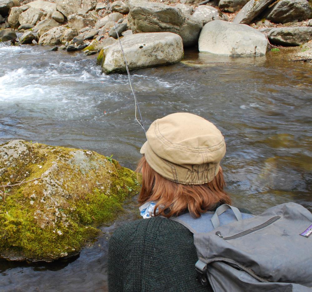 Fly fishing angler holding a fly rod fishing a small mountain creek fishing for brook trout.  Shen is leaning over a rock, wearing a sweater and a hat and backpack 